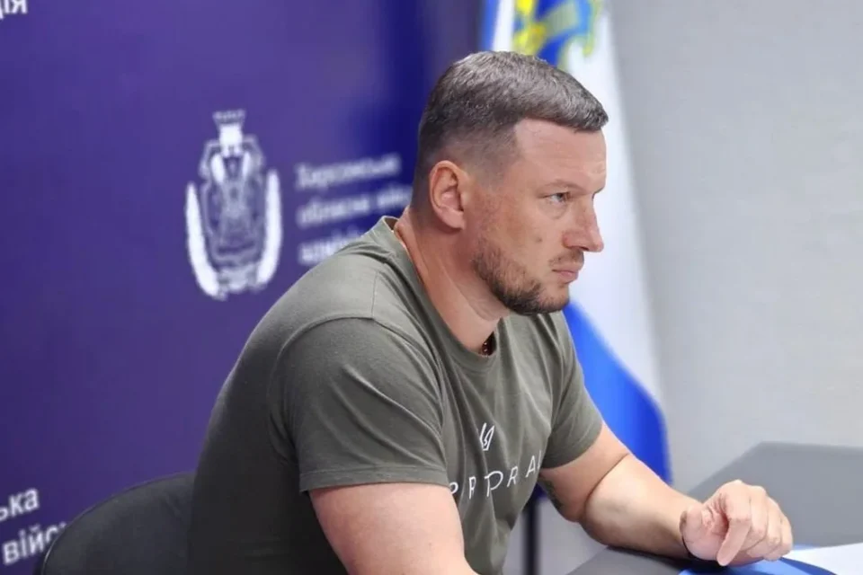 Чоловік у оливковій футболці сидить за столом на тлі банера Херсонської ОВА
