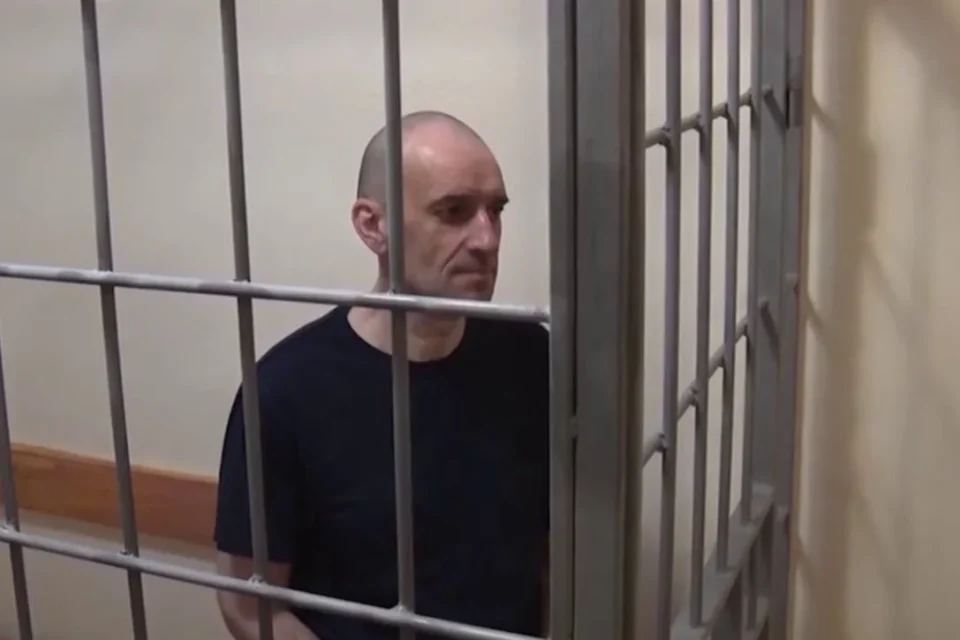 Лисий чоловік у чорній футболці стоїть за металевими ґратами